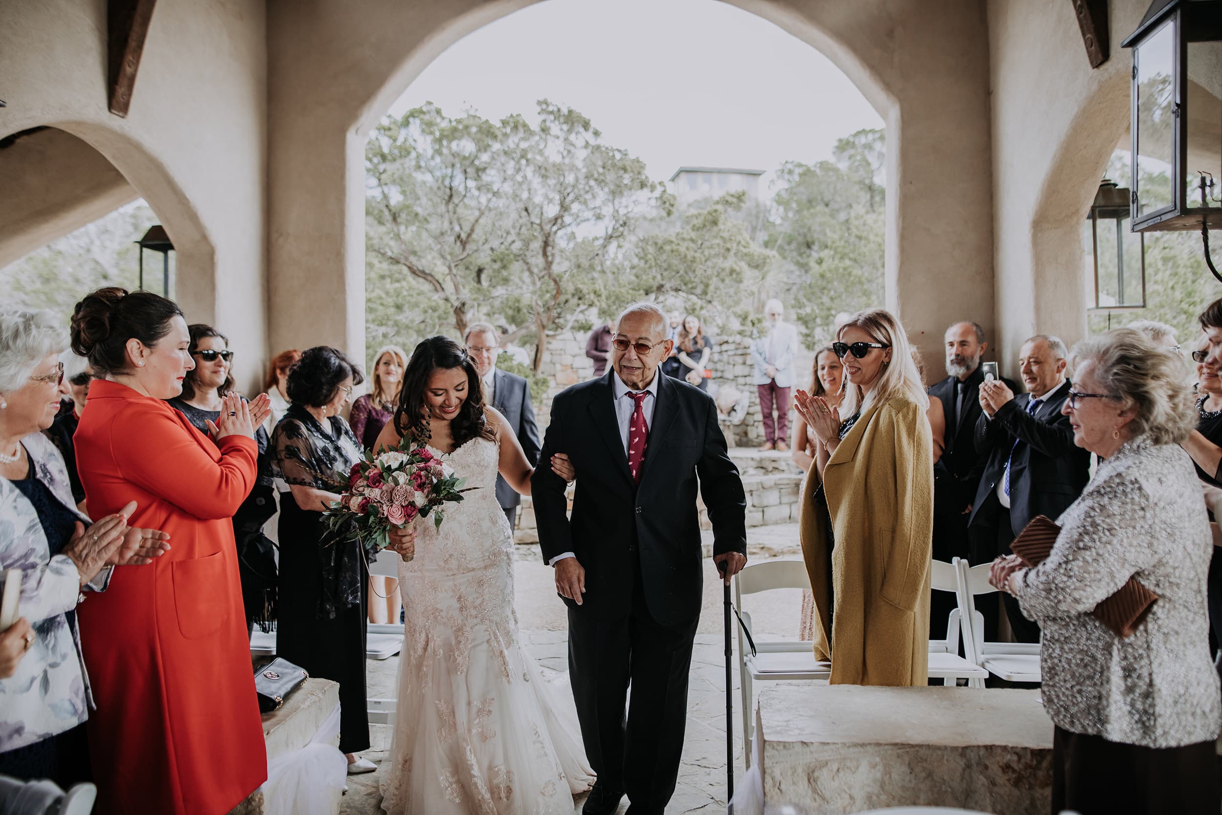 An older man walking a bride down the aisle as wedding guests applaud.