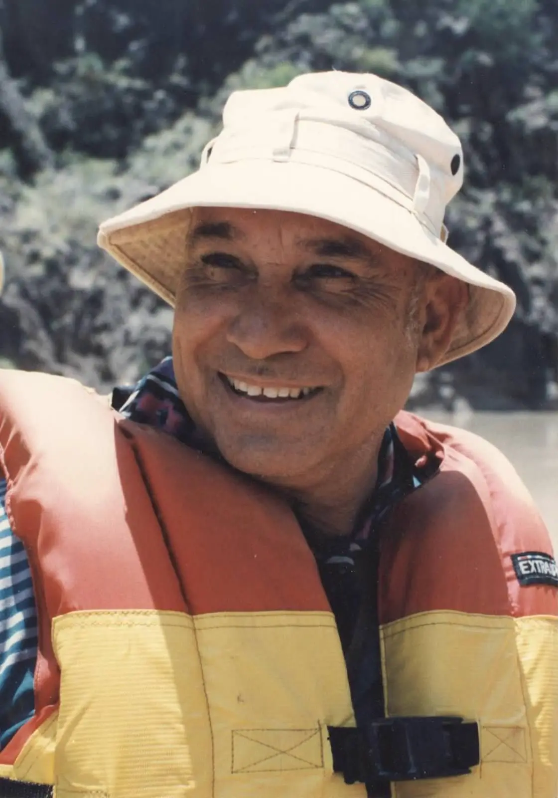 A smiling older man in a bucket hat and life jacket on the water