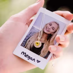Hand holding a Polaroid photo of a smiling blonde woman named Megan with a gold pendant necklace placed over the photo.