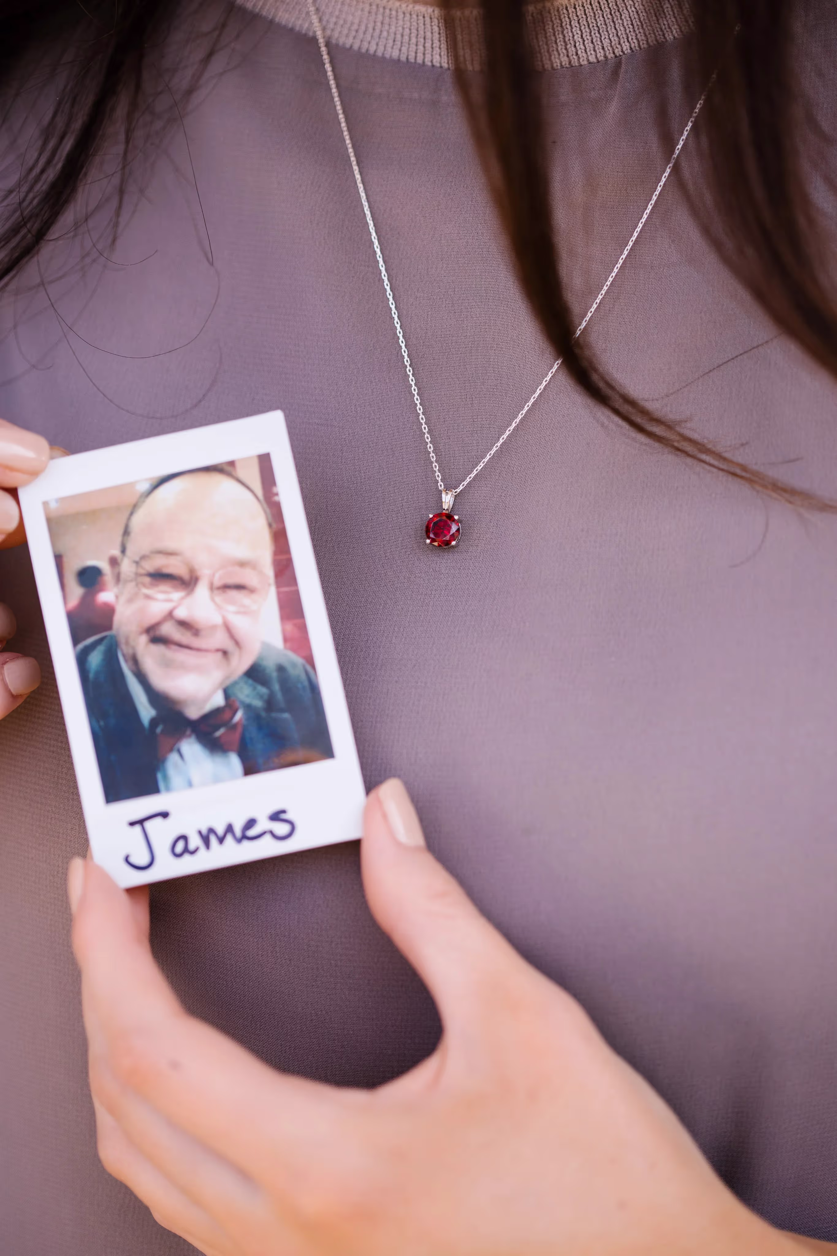 Close-up of a person wearing a silver necklace with a red gemstone pendant, holding a Polaroid photo labeled 'James' showing a smiling elderly man in glasses and a bow tie.