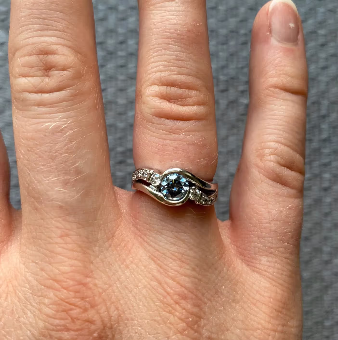 Hand wearing a silver ring with a central round blue gemstone and small clear stones on the band.
