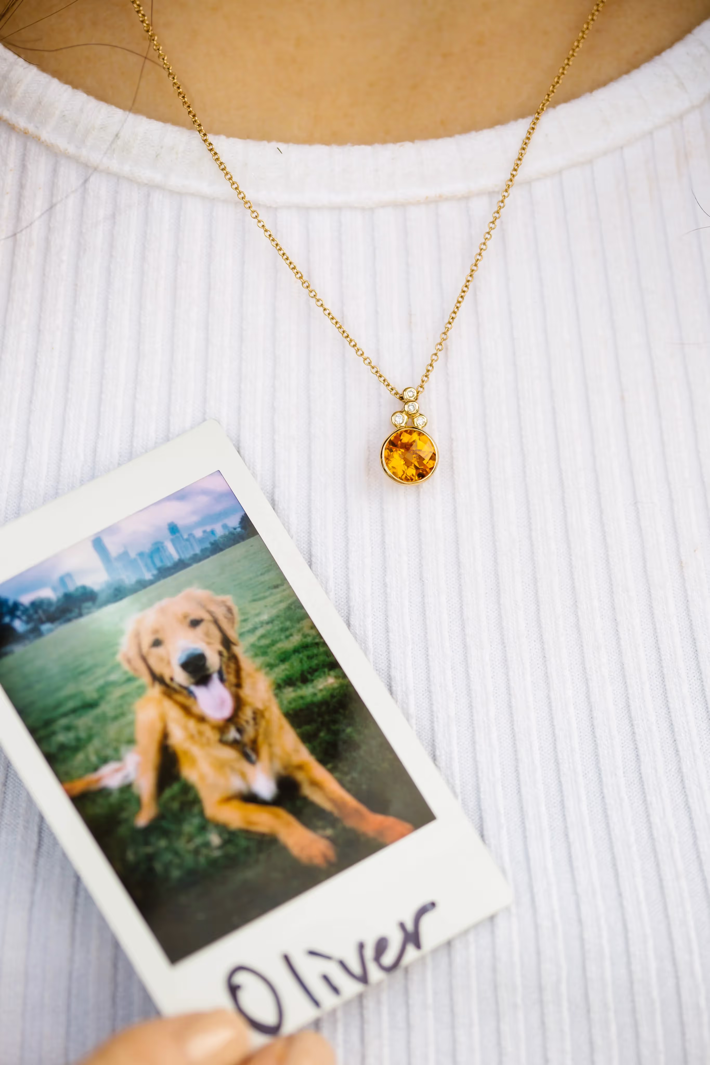 Person wearing a white ribbed shirt holding a Polaroid photo of a golden retriever dog named Oliver with a city skyline in the background, and a gold necklace with an orange gemstone pendant.