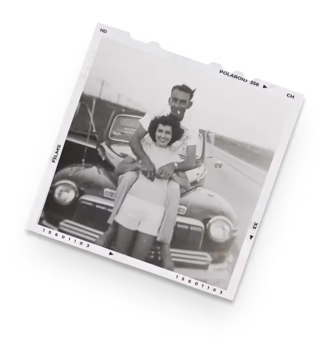 A black-and-white polaroid of a smiling couple in front of a vintage car.