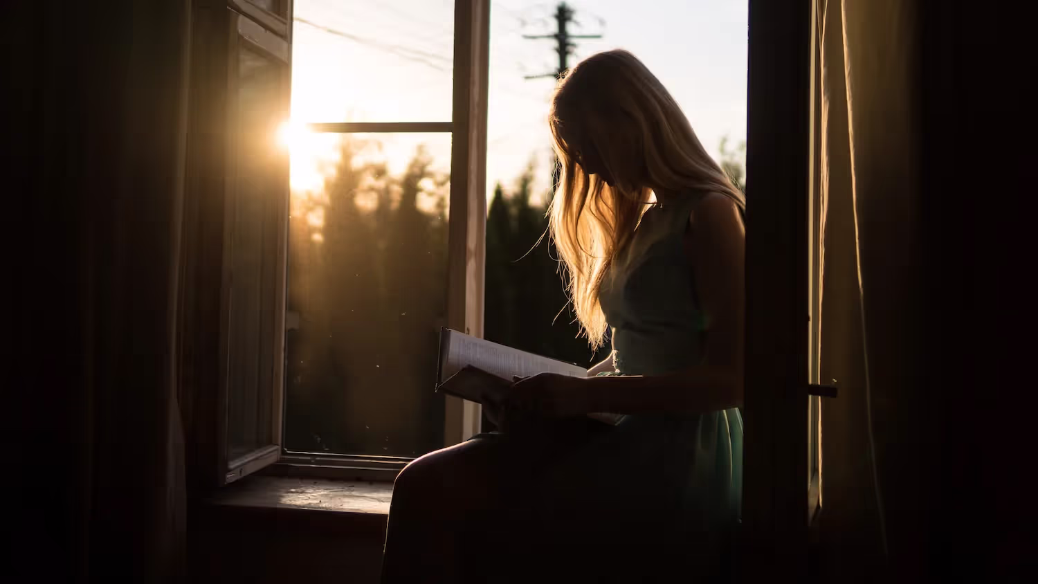 A woman sitting in a window sill, reading a book.