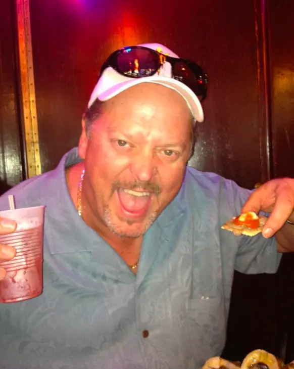 Rudy laughs with sunglasses on his cap, holding a drink and a slice of pizza.