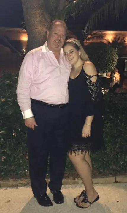 Rudy stands arm in arm with one of his daughters outdoors at night.