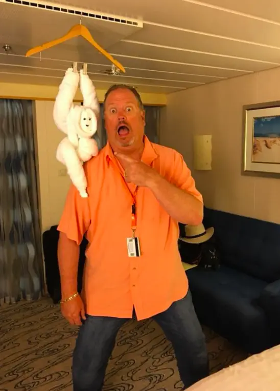 Rudy strikes a playful surprised pose in a cruise cabin with a towel monkey hanging behind him.