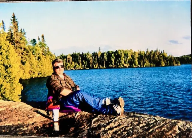 Steve relaxing in a camp chair on a rocky lakeshore lined with evergreen trees.