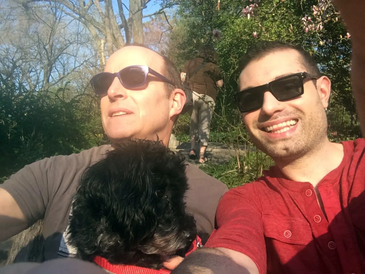 Marco and Jon in sunglasses taking a sunny selfie with their black dog.