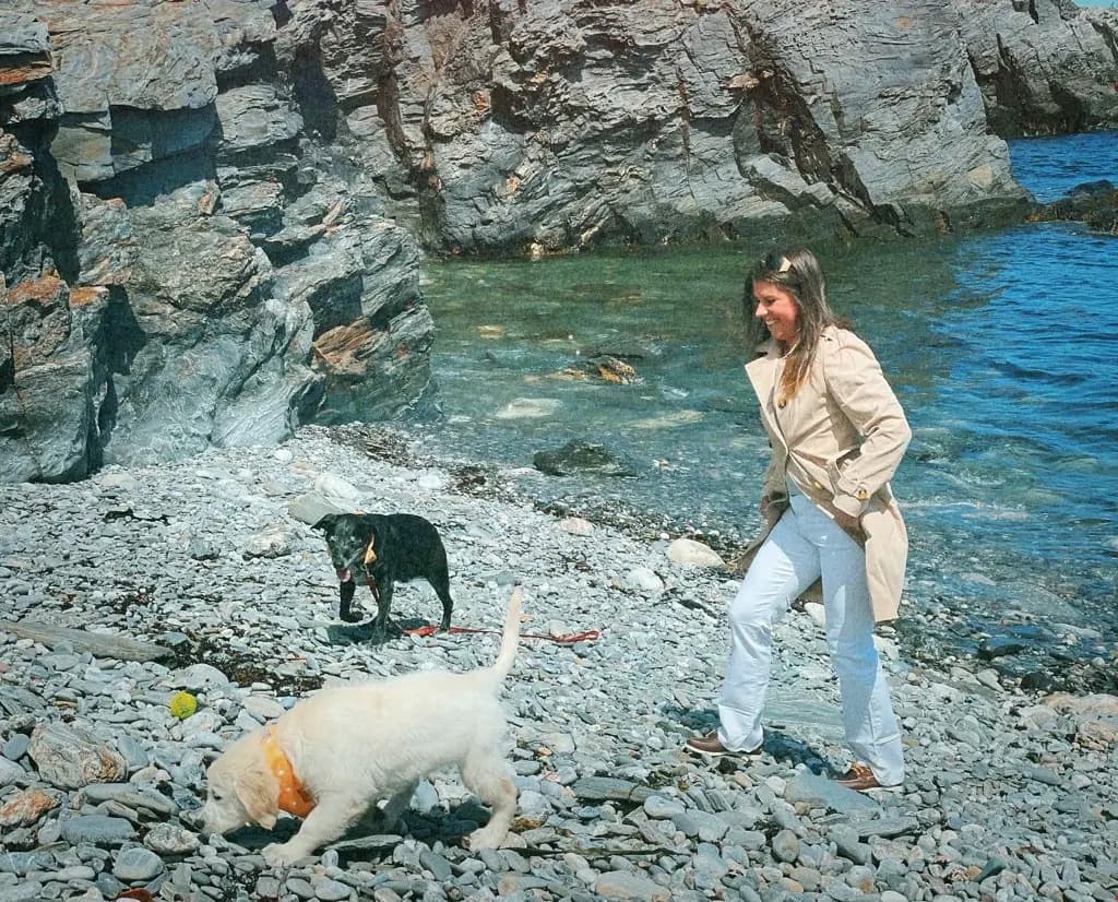 Lizzie recreating her mom's beach photo, walking a rocky shore in a trench coat with two dogs.