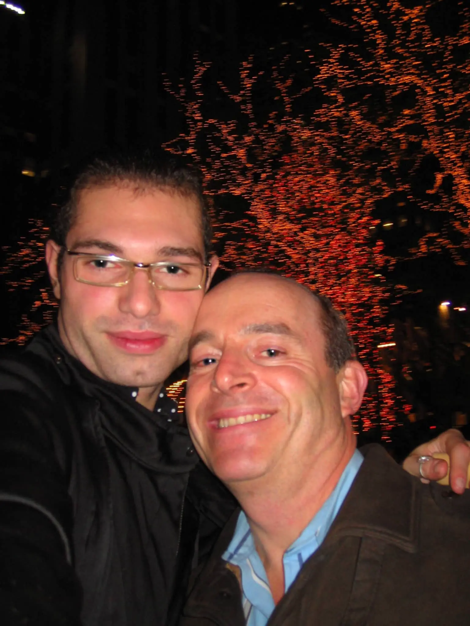 Marco and Jon cheek to cheek in front of a tree lit with red lights at night.
