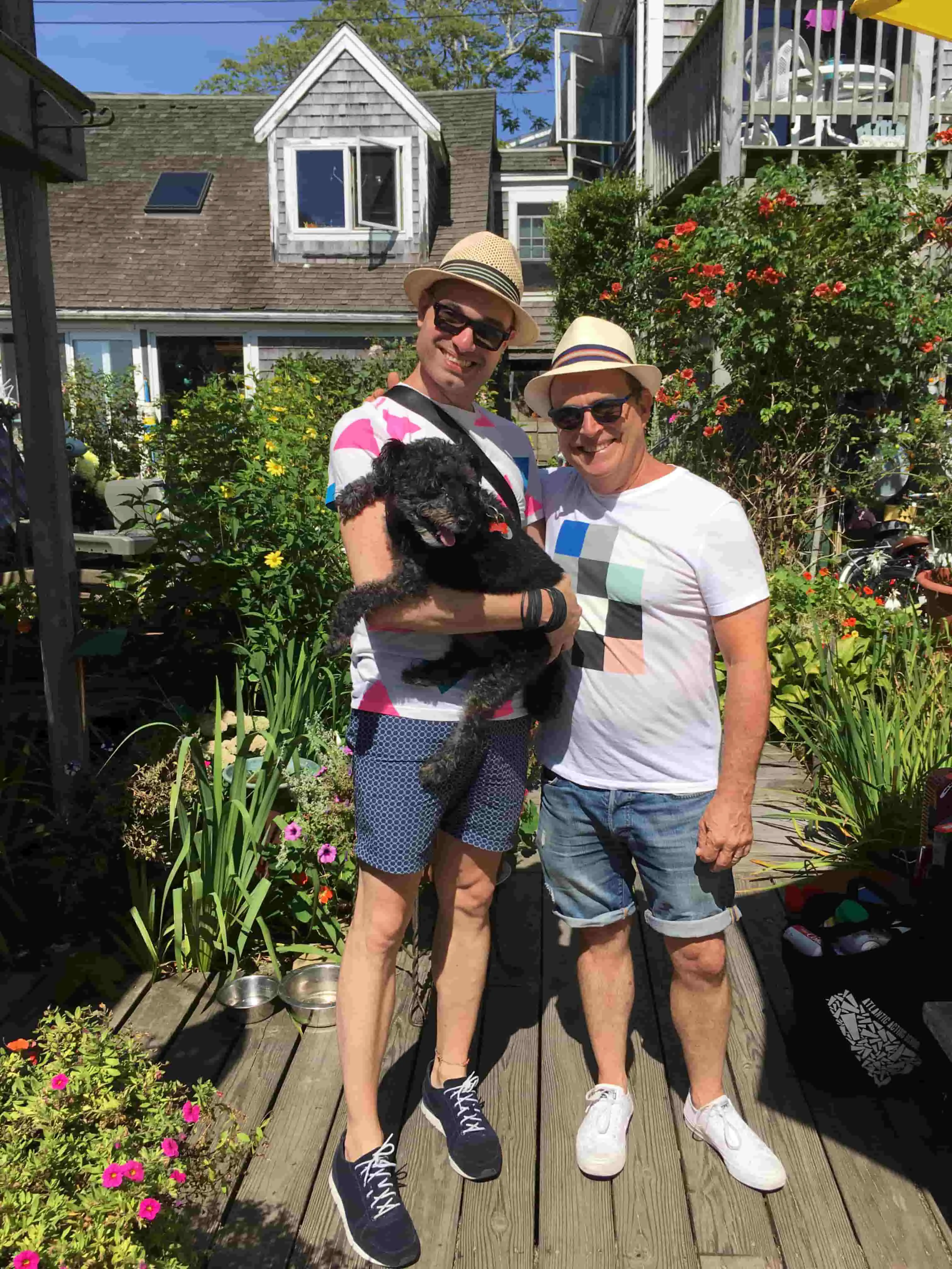 Two men in straw hats on a sunny garden deck, one holding a black dog.