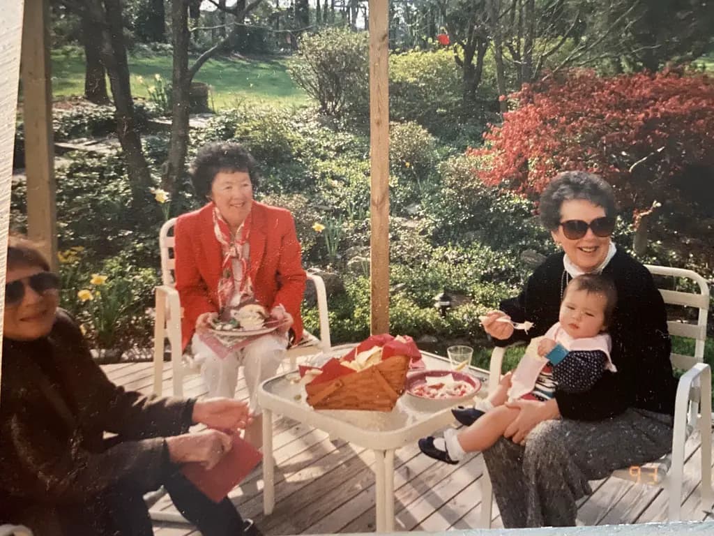 Three women sharing a meal on a garden patio, one holding a baby on her lap.