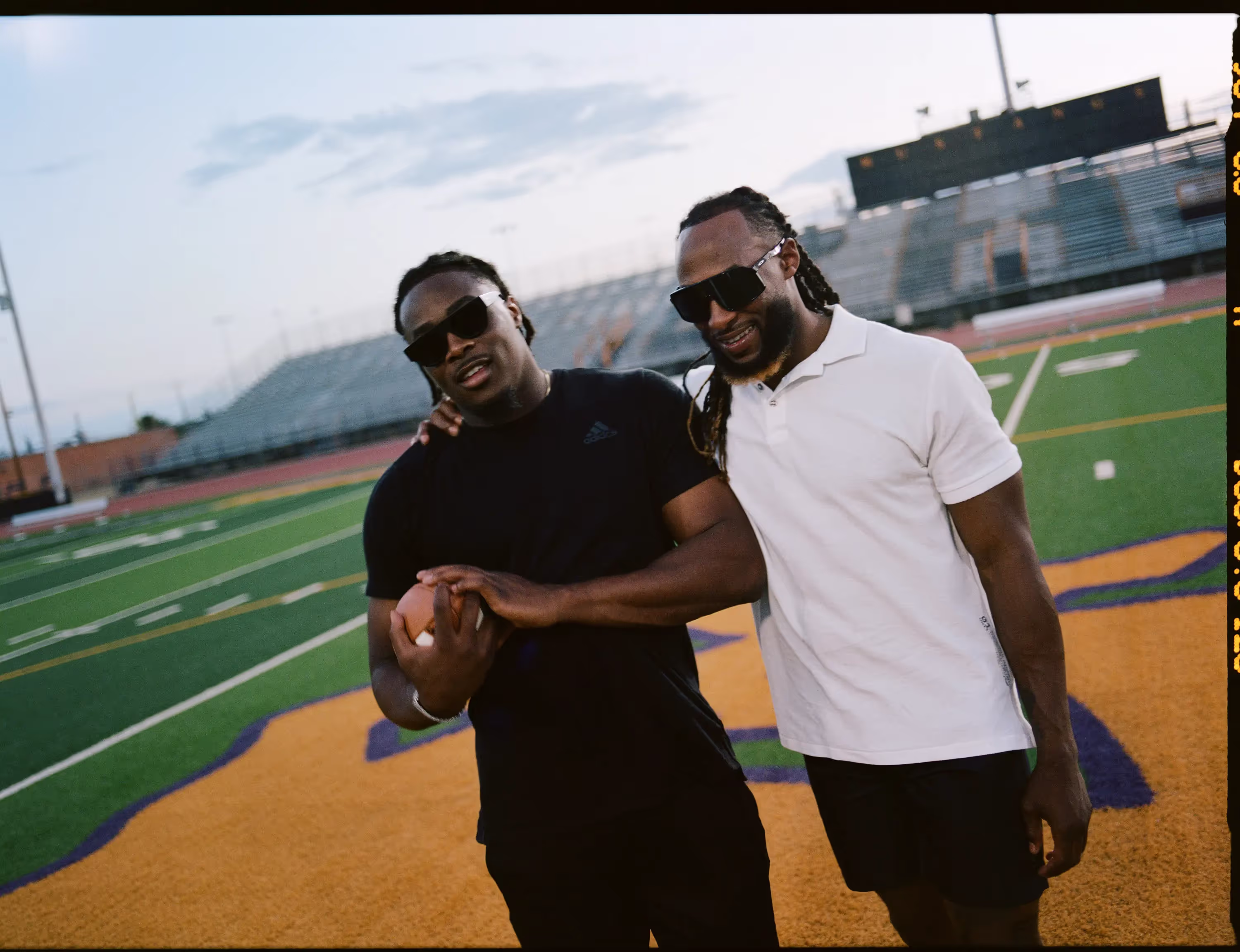 Two men pose together on a football field at dusk.