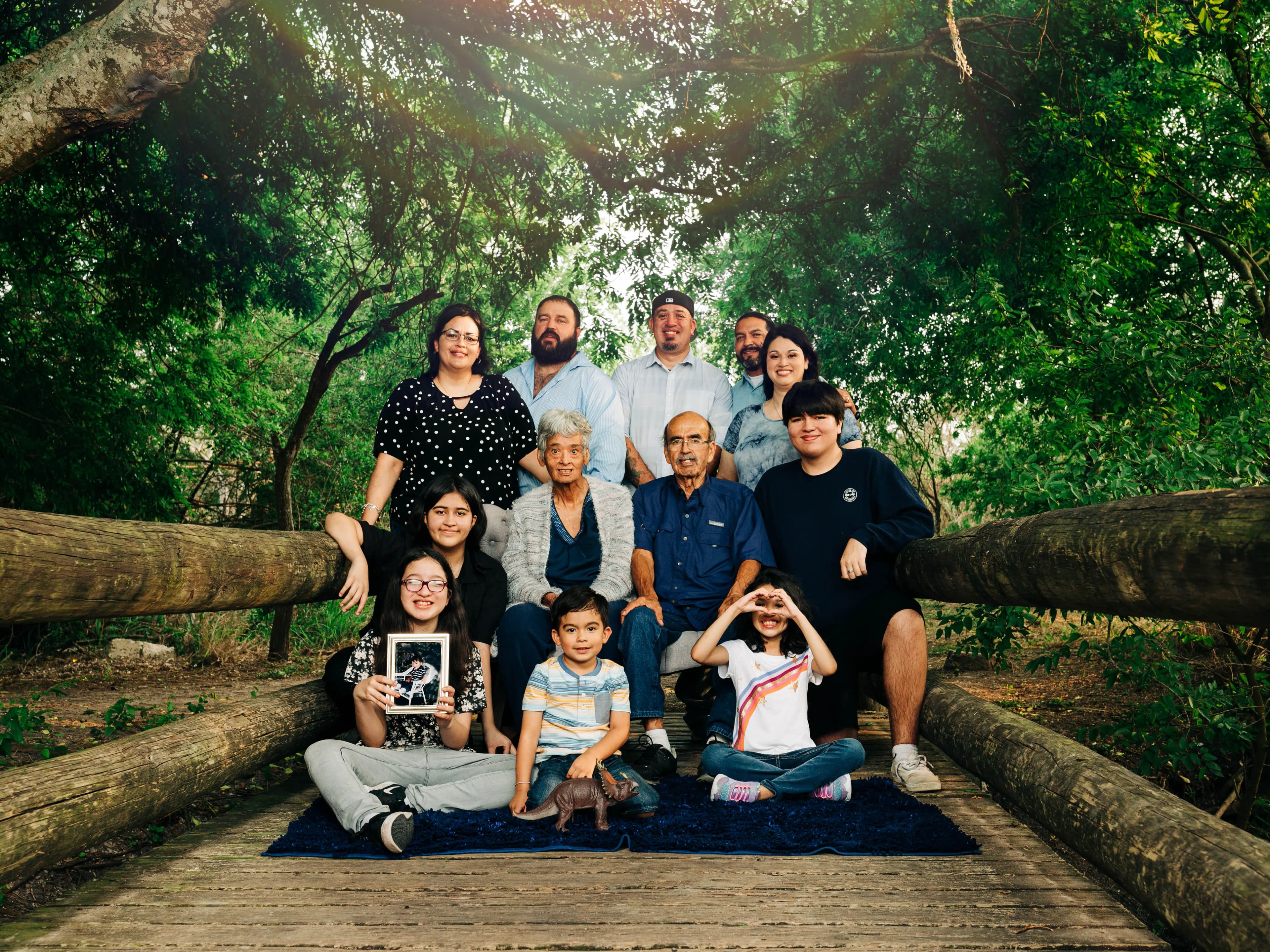 Large family portrait outdoors under trees, one person holding a framed photo.