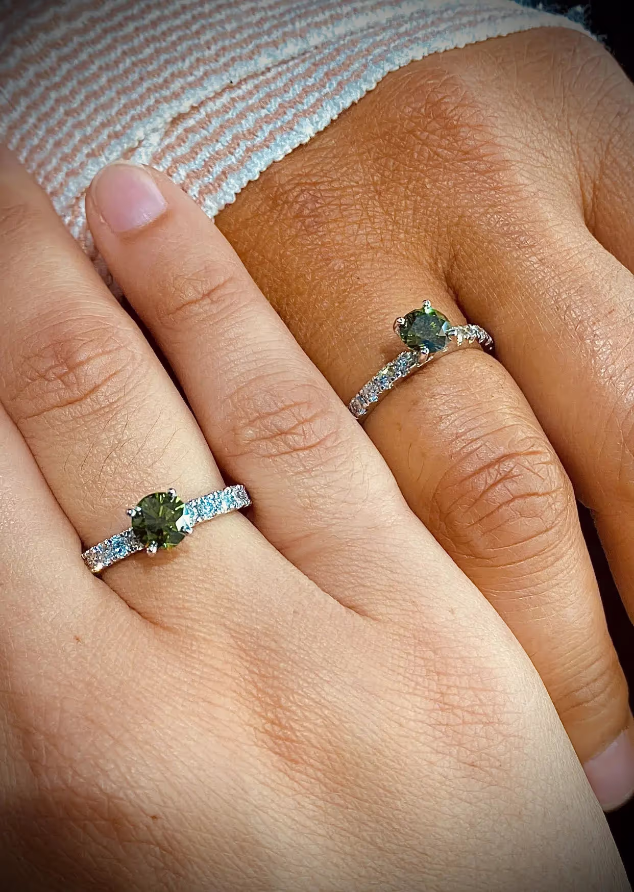Two hands side by side, each wearing a ring with a green memorial diamond.