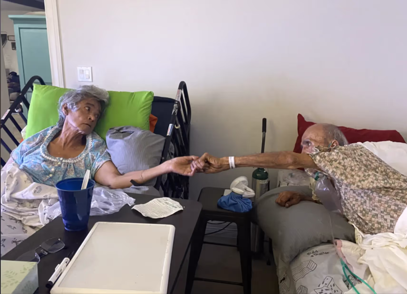 Rosa and Benny holding hands across their hospital beds.