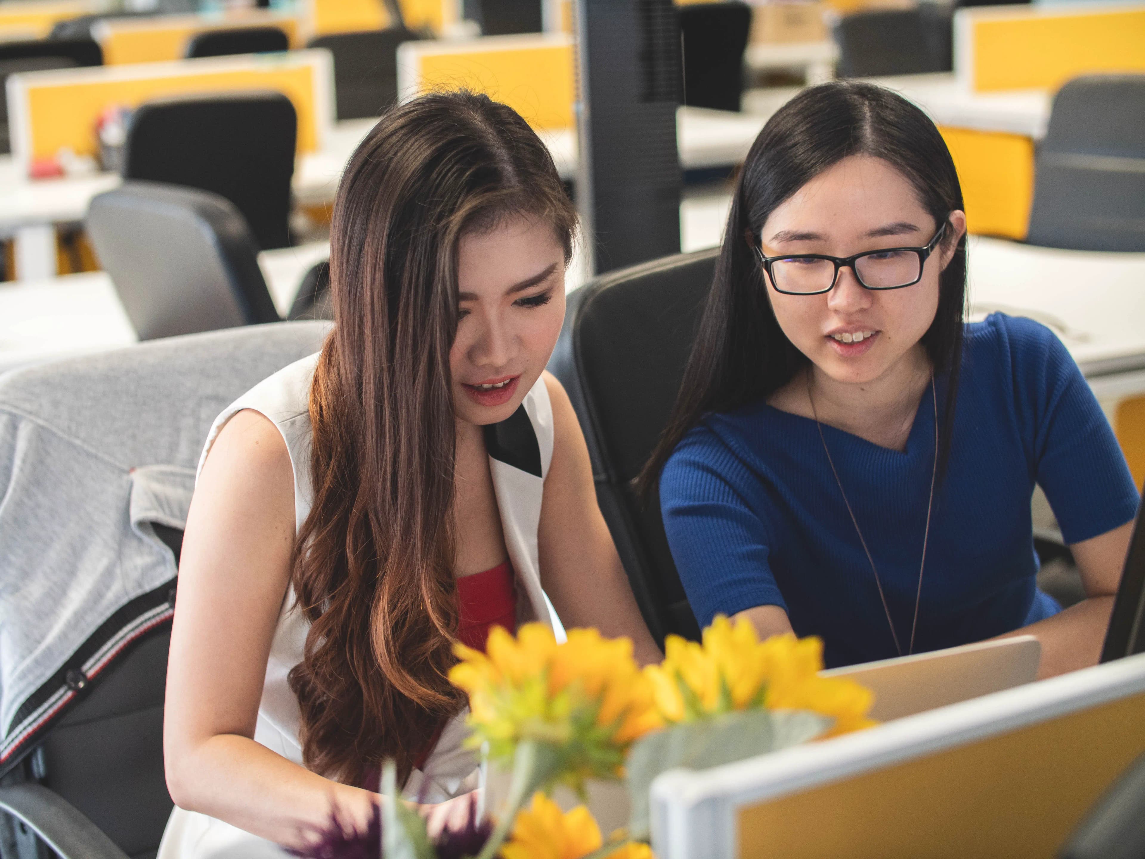 Two coworkers at a shared office desk look at a laptop together.