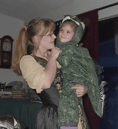 Woman in a costume holding a young child dressed in a green dragon costume.