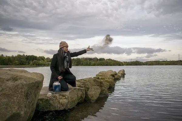 Person crouched on lakeside rocks tossing ashes into the wind over the water.