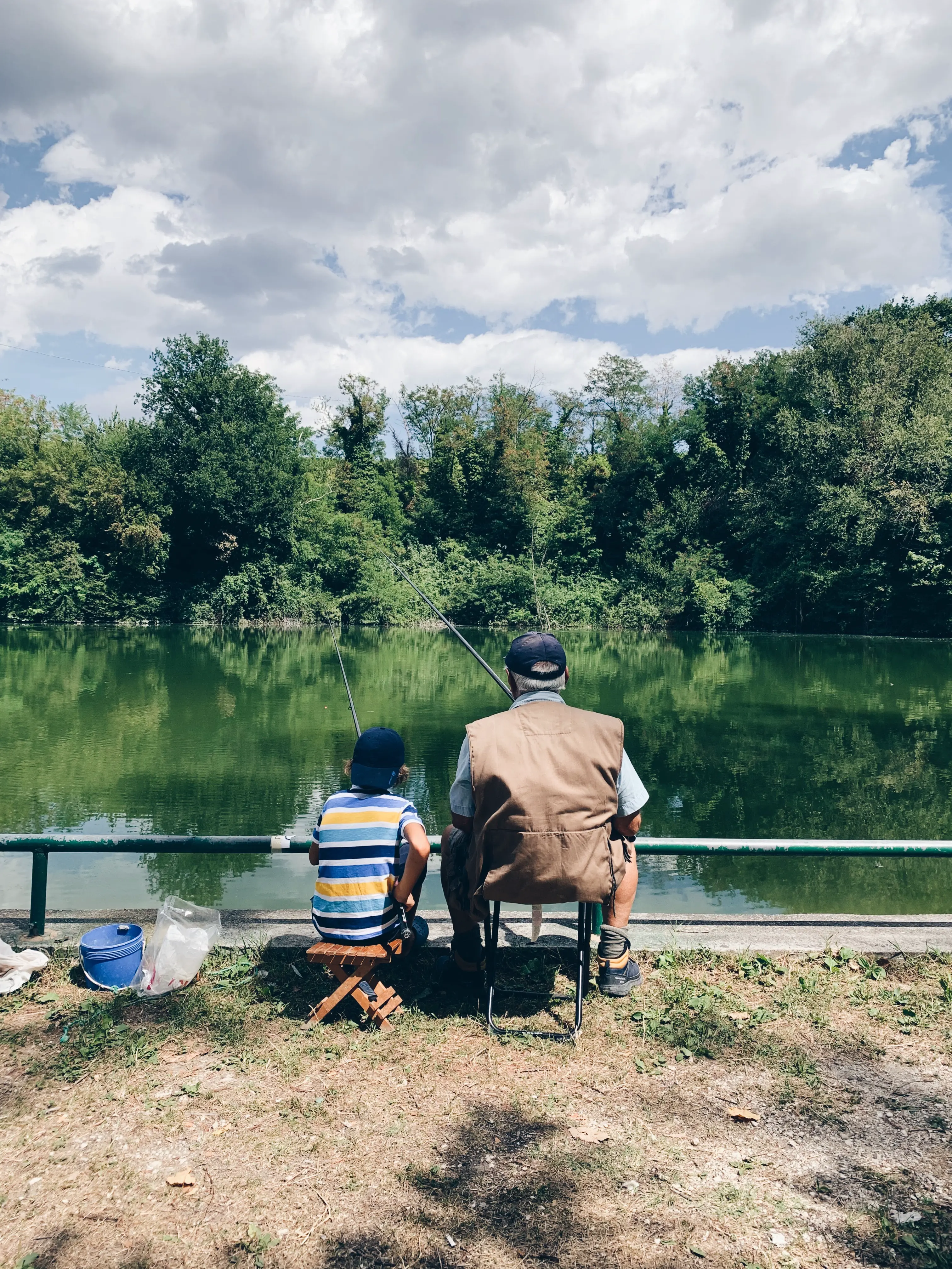 An older man and a young boy sit side by side fishing at a lake.