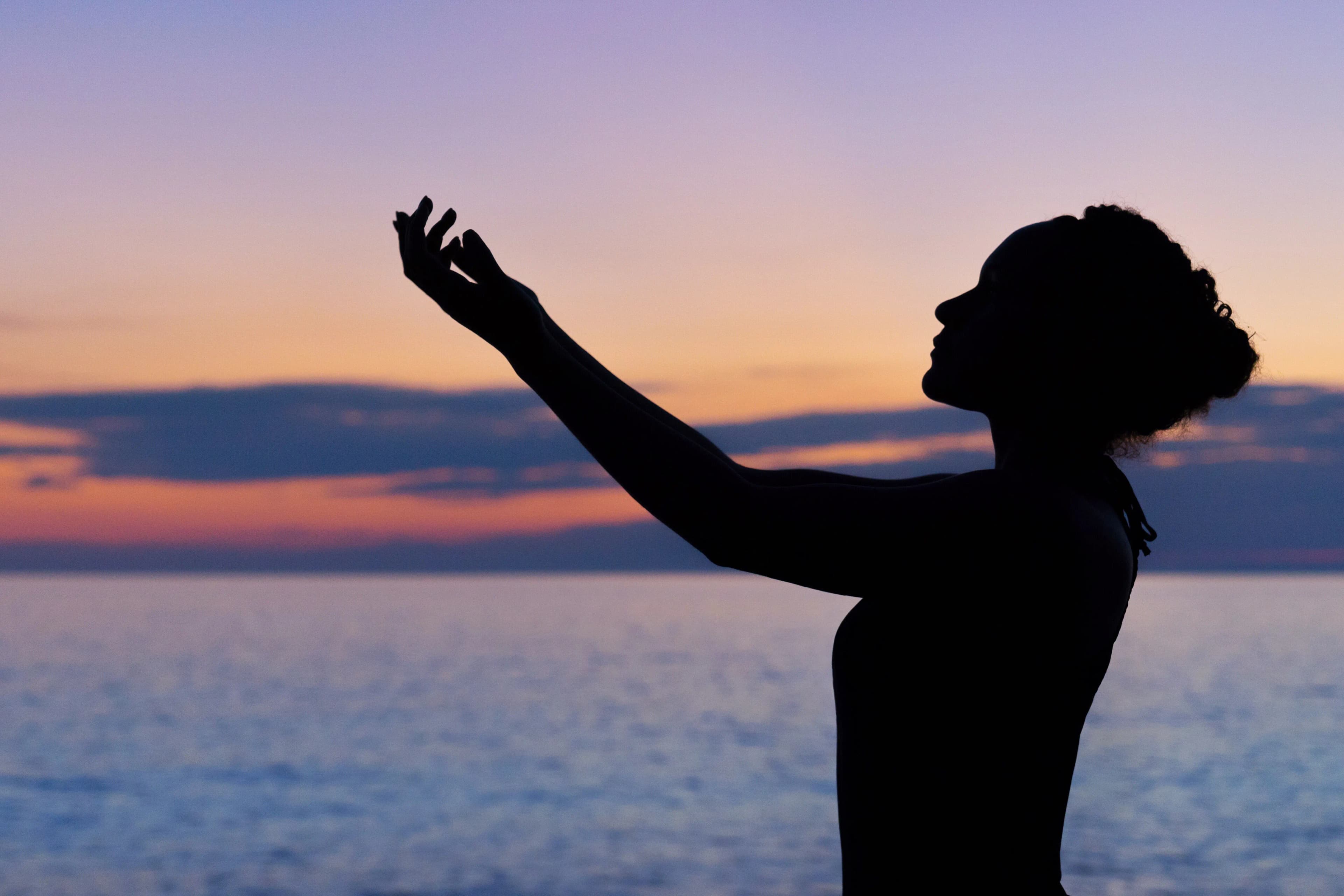 A woman's silhouette raises one arm toward a pastel sunset over the sea.
