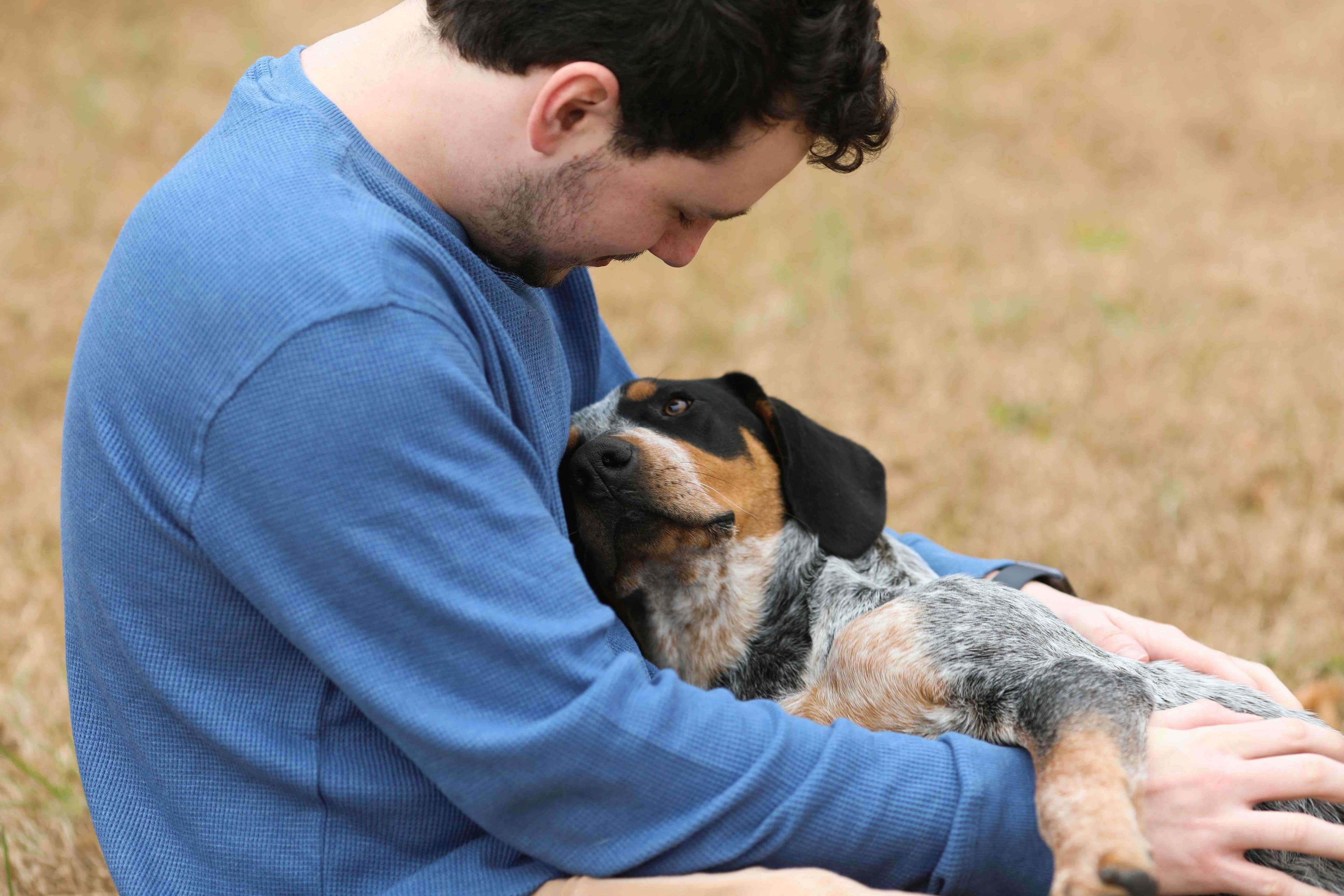 A man in a blue sweater cradling a senior dog in his lap in a grassy field