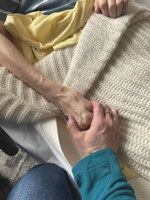 Two hands clasped gently over a knit blanket, one in a teal sleeve, in a polaroid print